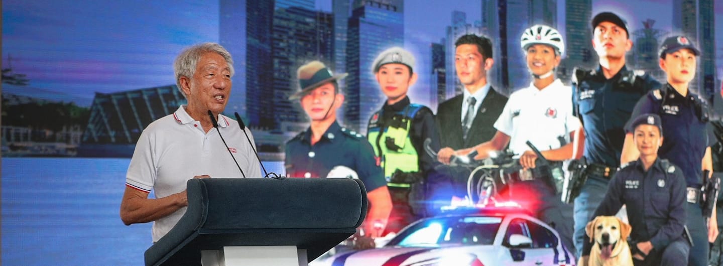 Man at podium in front of Singapore Police Force backdrop with officers, police car, and dog.